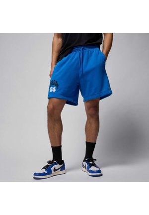 Persoon draagt felblauwe Jordan-short met bedrukking "84", zwarte sokken en witte, blauwe en zwarte Nike-sneakers, staat met handen in de zakken.