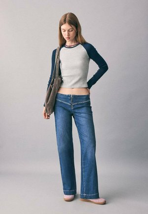 Mujer joven con un top corto de manga larga azul, jeans azules con detalles en cremallera, zapatos rosas y un bolso marrón, de pie frente a un fondo liso.