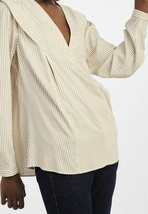 Personne portant un chemisier à rayures verticales beige et blanc à manches longues et un jean bleu foncé, visible du cou aux hanches.