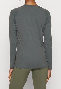 Koszula z długim rękawem w ciemnoszarym kolorze, o gładkiej teksturze, zaokrąglonym dekolcie i rękawach raglanowych. Zestawiona z oliwkowymi legginsami.