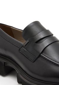 Des mocassins en cuir noir avec un bout arrondi, présentant une texture lisse, une large sangle sur le dessus, et une semelle noire robustes.