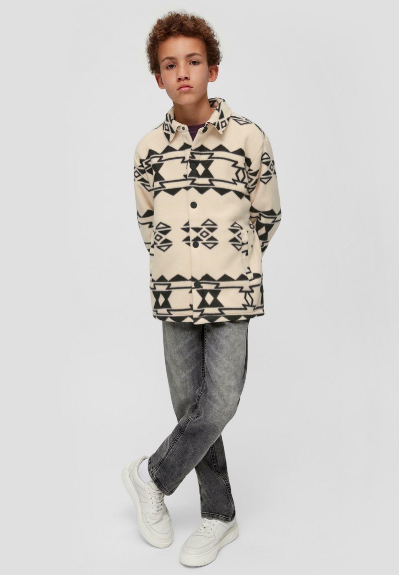 Fleece-Übershirt mit einem cremefarbenen Grundton und schwarzen geometrischen Mustern. Kombiniert mit grauen Jeans und weißen Sneakers. Mit Frontknopfverschluss.