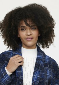 Geruite jas in blauwtinten met een textuur van wolmix. Crème gebreide trui eronder heeft verticale ribbelingen en subtiele details.