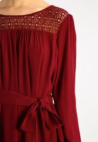 Punainen mekko, jossa on pitkät hihat, neliönmuotoinen kaula-aukko pitsiyksityiskohdilla ja sidottava vyö kehon muotoiluun. Pehmeä, sileä rakenne.