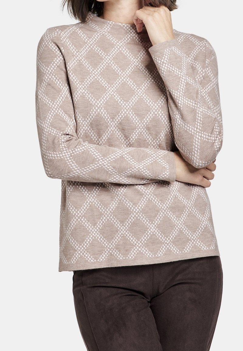 Femme portant un pull beige avec un motif losange blanc et un pantalon marron foncé, debout, un bras croisé et l'autre main près du menton.