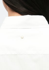 Λευκό πουκάμισο με γιακά, που διαθέτει έναν χρυσό κυκλικό κουμπί στο πίσω μέρος του λαιμού, με λεία υφή και κομψό σχέδιο.