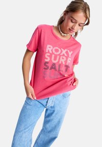 Lys pink t-shirt med teksten "ROXY SURF SALT SUN" trykt i varierende bredder og farver. Korte ærmer, afslappet pasform, blødt stof, parret med lyseblå jeans.