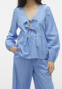Light blue, long-sleeve top with a deep V-neck, tie detail at the bust, and a peplum hem. Vertical white stripes and textured fabric.