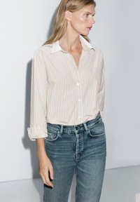 Hemd mit Kragen und langen Ärmeln in Cremefarbe mit vertikalen lavendelfarbenen und marineblauen Streifen. Dazu trägt man hochgeschnittene blaue Jeans. Lässiges Design und Passform.