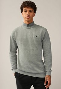 Un pull en tricot gris présente un design texturé, des poignets côtelés et un col rond. Un petit logo brodé est situé sur la poitrine.