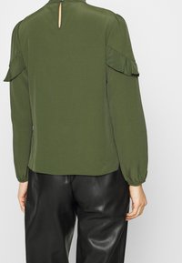 Olivgrön långärmad blus med rynkade axelapplikationer och öppning med nyckelhål bak. Slät, lättviktig tyg. Kombinerad med svarta byxor.