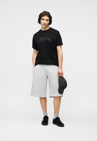 Νεαρό άτομο με μαύρο γραφικό μπλουζάκι, ανοιχτό γκρι σορτς μέχρι το γόνατο, γκρι κάλτσες, μαύρα αθλητικά παπούτσια, κρατώντας ένα σκούρο καπέλο στο δεξί χέρι.