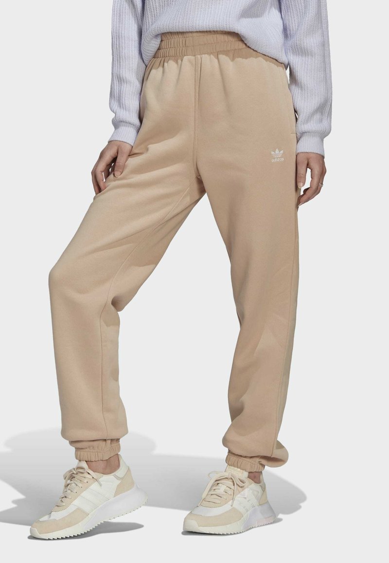 Personne portant un pantalon de survêtement Adidas beige, des baskets blanches et un pull gris clair, debout les mains le long du corps devant un fond uni.