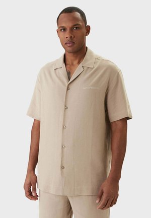Uomo che indossa una camicia beige a maniche corte con bottoni e pantaloni abbinati, in piedi davanti a uno sfondo chiaro e uniforme.