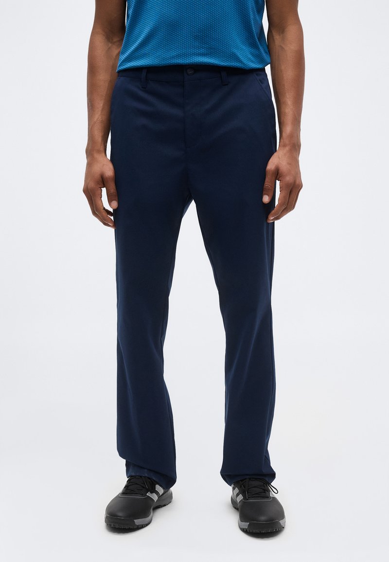 Άνδρας που φοράει σκούρο μπλε παντελόνι, μαύρα αθλητικά παπούτσια και μπλε υφασμένο κοντομάνικο πουκάμισο, στέκεται σε απλό φωτεινό φόντο.