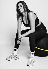 Zwarte sportbeha met gele accenten, zwarte legging met witte strepen, en grijze sportschoenen. Zittend op een zwarte fitnessbal.