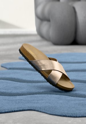 Ένα μεταλλικό σανδάλι slip-on με σταυρωτούς ιμάντες πάνω σε υφασμάτινο μπλε χαλί, με απαλό γκρι έπιπλο στο φόντο θολωμένο.