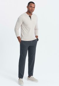 Hombre de pie con las manos en los bolsillos, vistiendo una camiseta henley de manga larga beige, pantalones gris oscuro, zapatillas beige y un reloj de pulsera.