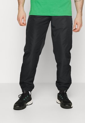 Man draagt zwarte taps toelopende broek met elastische boorden, zwarte sneakers met groene accenten en een groen shirt, staand op een lichtgrijze vloer.