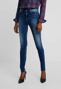 Jeans ajustados de mezclilla en azul, con un lavado desgastado, bolsillos frontales y cinturilla, combinados con tacones negros de punta y una camisa de cuadros.