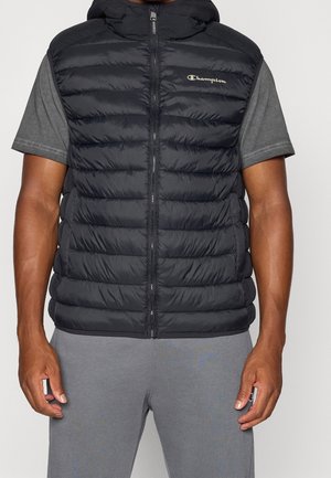 Homme portant une doudoune noire sans manches avec une fermeture éclair et le logo "Champion" sur un t-shirt gris à manches courtes et un pantalon gris.