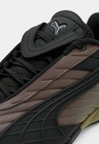 Prim-plan al unei tenisi Puma negre și maronii, cu un panou lateral texturat, șireturi negre și logo-ul Puma pe limbă.