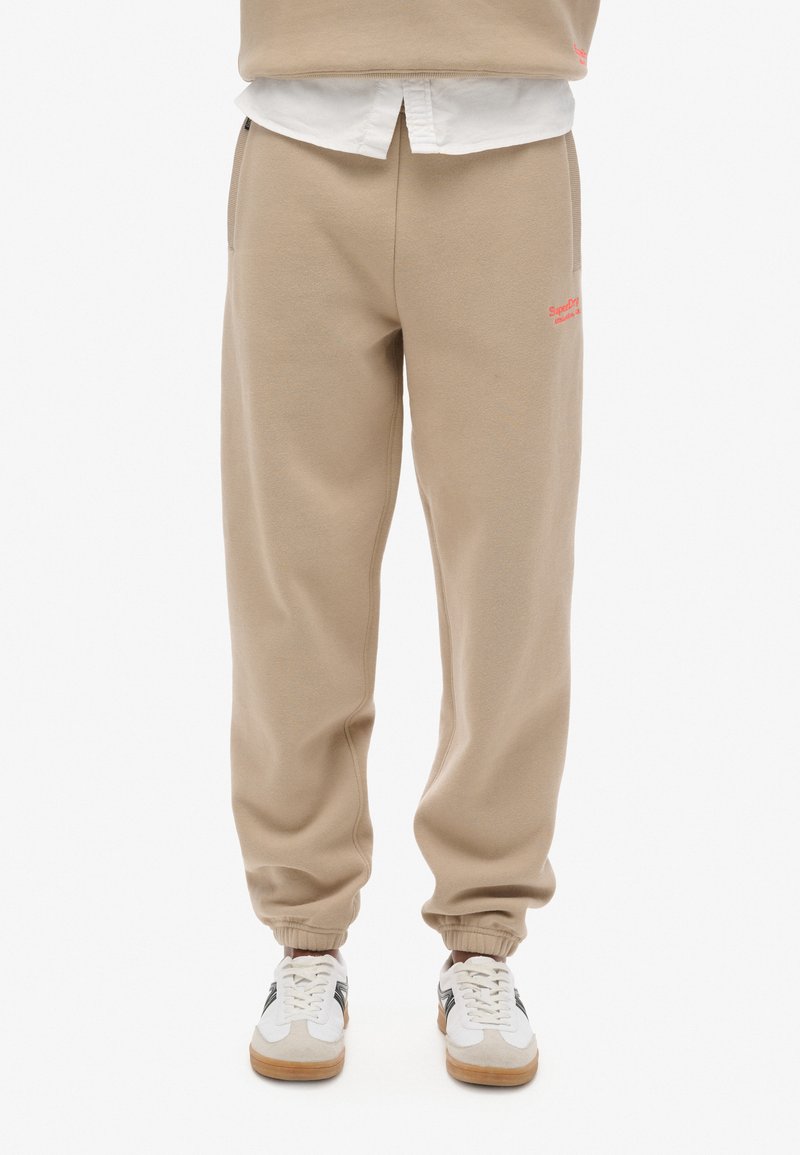 Pantalones de chándal beige confeccionados en suave algodón, con una cinturilla elástica, puños acanalados y bolsillos laterales, con un pequeño logo rojo en el muslo.