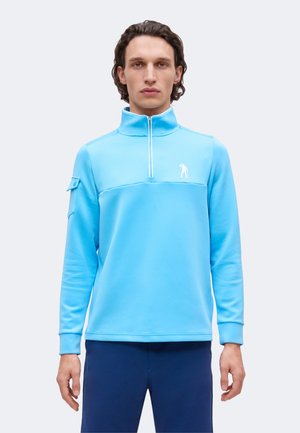 Homme portant un pull léger bleu clair à fermeture éclair quart de zip avec un petit logo blanc sur la poitrine et un pantalon bleu marine, debout devant un fond uni.