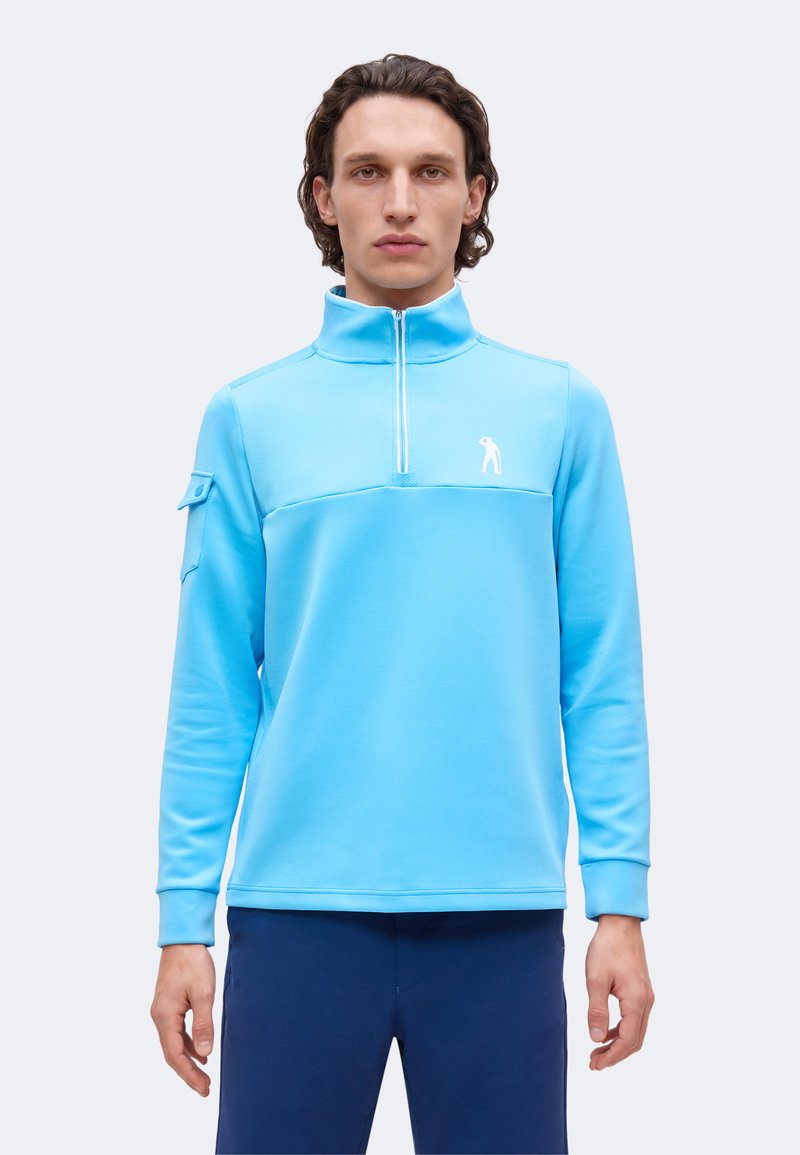 Homme portant un pull léger bleu clair à fermeture éclair quart de zip avec un petit logo blanc sur la poitrine et un pantalon bleu marine, debout devant un fond uni.
