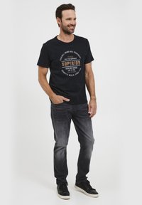 Camisa negra de cuello redondo con estampado gráfico en marrón y blanco, combinada con jeans gris oscuro y zapatillas negras, posando frente a un fondo blanco.