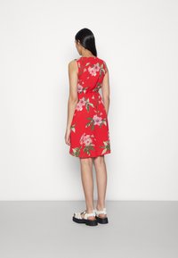 Vestido floral vermelho com design sem mangas, cintura com cinto e tecido estampado. Usado com sandálias brancas de tiras e sola preta grossa.
