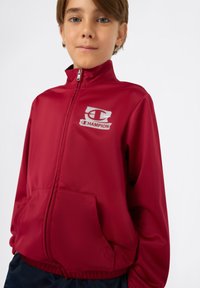 Rode zip-up jack met een gladde synthetische stof, voorzien van een wit "Champion" logo en zijzakken. Heeft een hoge kraag en elastische manchetten.