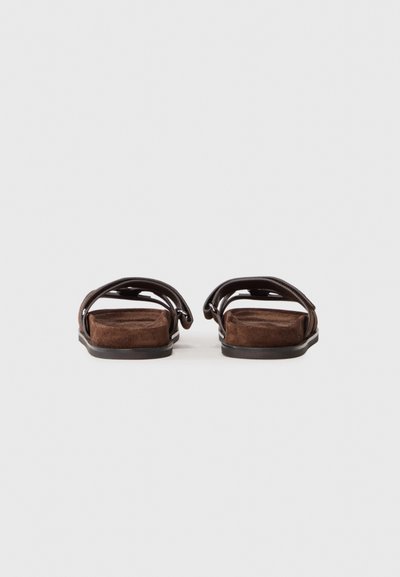 Brown suede sandals with a flat sole, featuring two wide straps and a smooth, minimalistic design. No visible hardware or patterns.