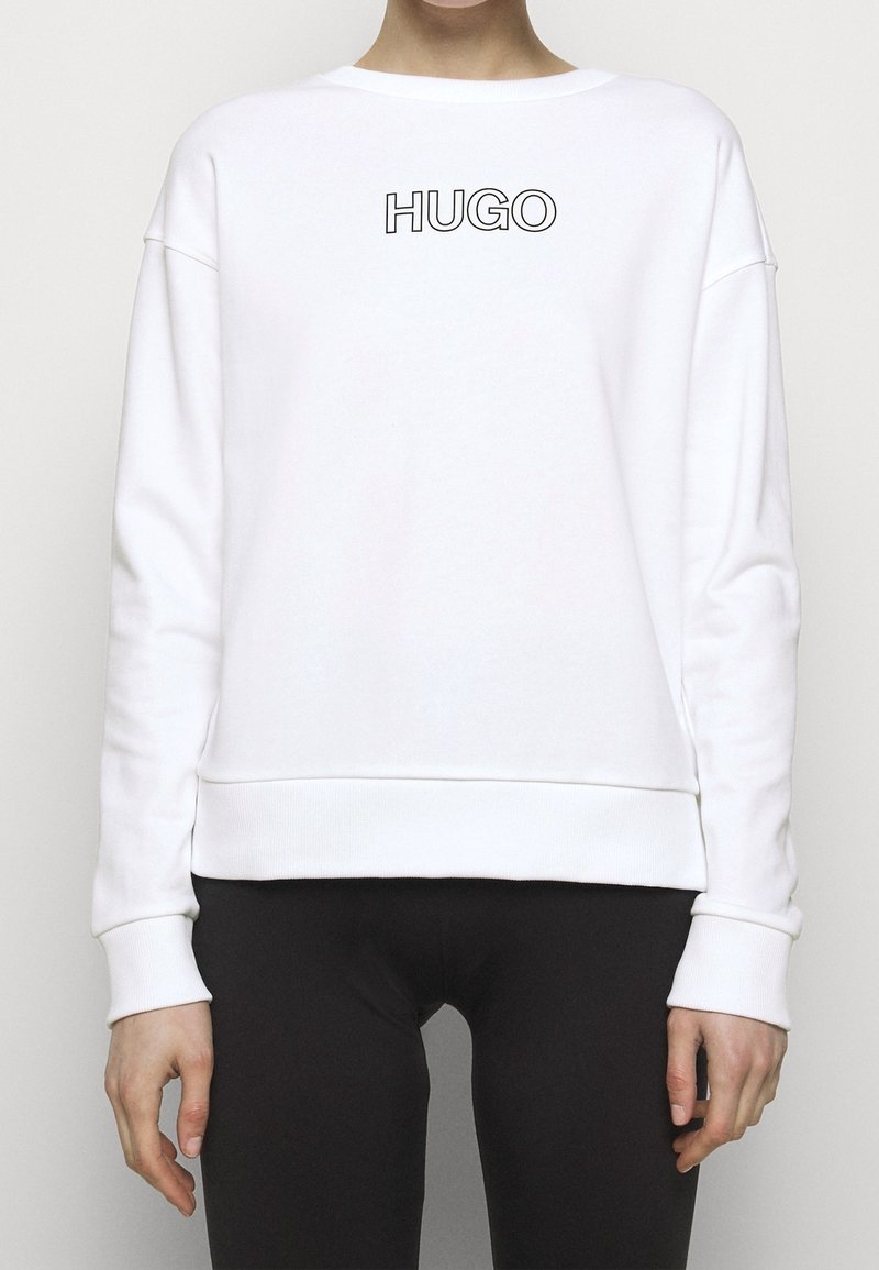 Personne portant un sweat-shirt blanc avec un texte noir "HUGO" sur la poitrine et un pantalon noir, debout devant un fond clair uni.