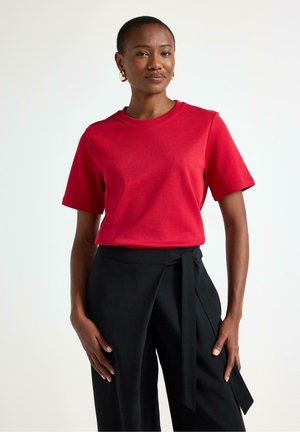 Punainen puuvillapaita lyhyillä hihoilla ja pyöreällä kaula-aukkolla, yhdistettynä mustiin housuihin, joissa on solmittava vyö. Sileä pinta, rento muotoilu.