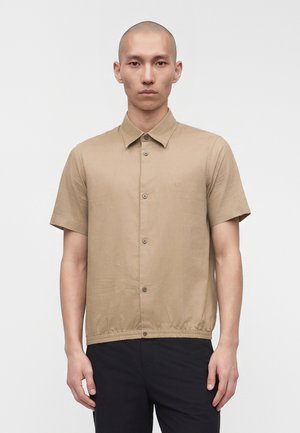 Mees habetuna, kandes beeži lühikeste varrukatega nööbitava särgi ja mustad püksid, seisab ühevärvilise valge tausta ees.