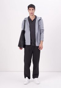 Sudadera gris con cremallera, camiseta oscura, pantalones negros y zapatillas blancas. Modelo sujetando una bolsa de mano negra. Diseño sencillo, look casual.