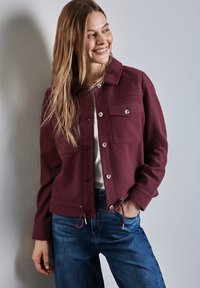 Bordeauxfarbene Jacke mit Kragen, zwei Brusttaschen, Druckknöpfen und Kordelzug in der Taille, kombiniert mit blauen Jeans.