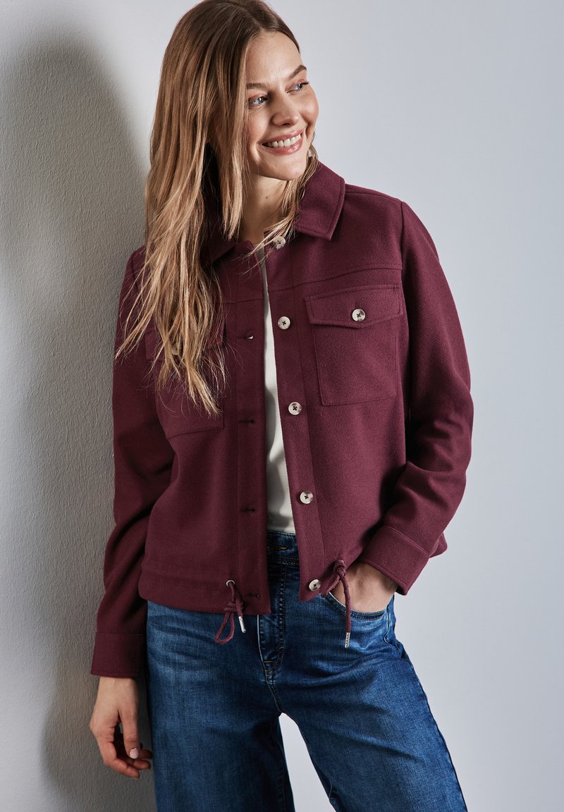 Bordeauxfarbene Jacke mit Kragen, zwei Brusttaschen, Druckknöpfen und Kordelzug in der Taille, kombiniert mit blauen Jeans.