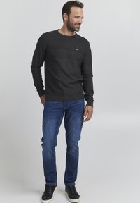 Mørk grå strikkegenser med teksturert mønster over brystet, ribbestrikkede mansjetter og en liten logo. Kombinert med blå denimjeans og svarte joggesko.
