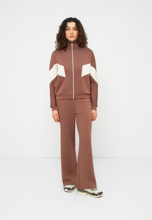 Moteris su garbanotais plaukais stovi, vilkinti rudą sportinį kostiumą su baltais chevronų raštais rankovėse ir dėvi šviesiai rudus sportinius batelius, fone yra vientisa balta siena.