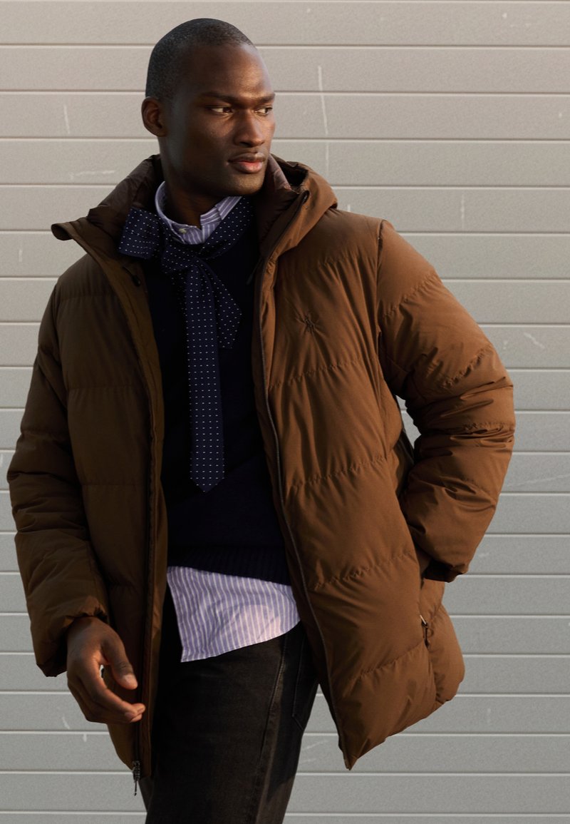 Brown puffer jacket featuring a hood, zip closure, and textured design. Worn over a navy sweater and light striped shirt with a polka-dot tie.