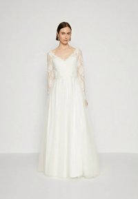 Sieviete, tērpusies baltā kāzu kleitā ar mežģīņu garajām piedurknēm un pilnu tūlļa svārku, stāv uz vienkārša baltā fona.