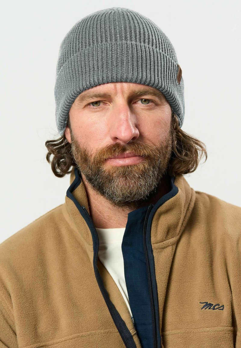 Homme avec barbe brune et cheveux bouclés portant un bonnet gris en tricot et une veste polaire beige avec une fermeture éclair bleu marine et le logo "mcs".