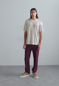 Camiseta de manga corta de un beige claro con texto en relieve, combinada con pantalones burdeos y zapatillas beige con acentos contrastantes.