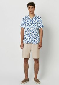 Kurzarmhemd mit blauem Blattmuster auf weißem Hintergrund, kombiniert mit hellbeigen Shorts und braunen Slippern.