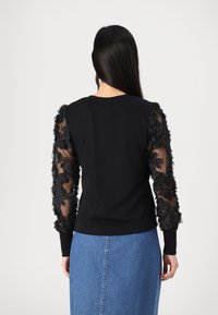 Schwarzer Pullover mit transparenten, strukturierten Ärmeln, verziert mit floralen Applikationen. Glatte Körperpartie mit Rundhalsausschnitt; kombiniert mit einem Jeansrock.