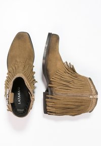 Brązowe zamszowe botki za kostkę z frędzlami, spiczastymi czubkami i bocznymi zamkami błyskawicznymi. Posiadają zdobienia w postaci przeszyć oraz niski, warstwowy obcas.