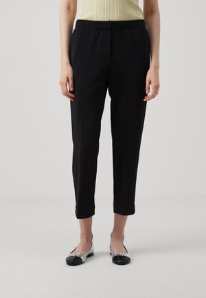 Personne portant un pantalon noir fuselé avec ourlets retroussés, un haut beige côtelé et des ballerines argentées à bouts noirs, debout devant un fond uni.