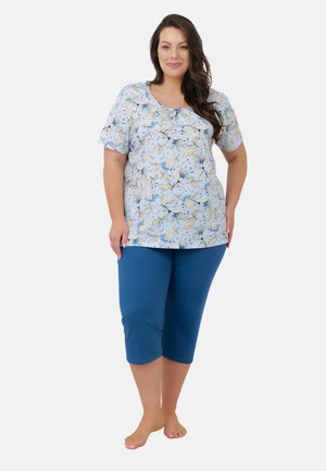 Kobieta stoi boso, ubrana w jasnoniebieską kwiatową bluzkę z krótkim rękawem i jednolite niebieskie spodnie za kolano, uśmiecha się, trzymając dłoń na biodrze.